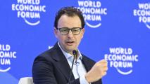A man in a suit and glasses speaks into a microphone at the World Economic Forum, with the event's logo visible in the background.