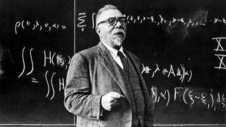 An older man in a suit stands in front of a chalkboard filled with mathematical equations, holding a piece of chalk.