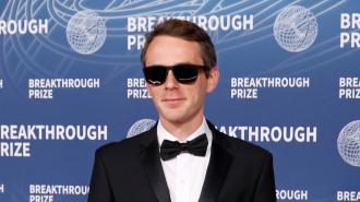 A man in a black tuxedo and sunglasses stands in front of a blue backdrop with the Breakthrough Prize logo and text.