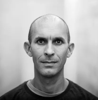 A man with a shaved head and dark eyes stares directly at the camera. The background is blurred and in grayscale.