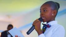 A young person in a school uniform speaks into a microphone while holding a piece of paper.