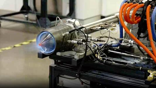 A close-up of an engine test firing, showcasing various connected hoses and cables, with the engine emitting blue flames from the nozzle, reminiscent of a powerful jet engine.