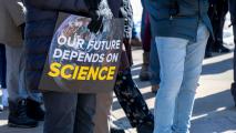A person wearing gloves holds a sign reading "OUR FUTURE DEPENDS ON SCIENCE" with an image of Earth; several people stand nearby on a sidewalk.