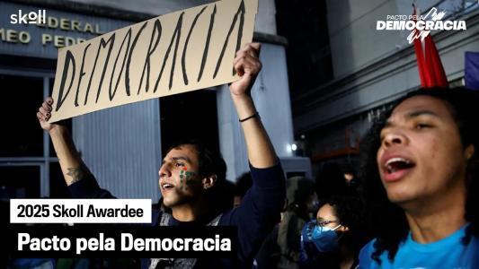 A group of people at a demonstration, one holding a large sign that reads "Democracia"; text overlay reads "2025 Skoll Awardee Pacto pela Democracia.