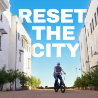 A person rides a bicycle between two modern white buildings under a clear blue sky, with large text reading "RESET THE CITY" above.