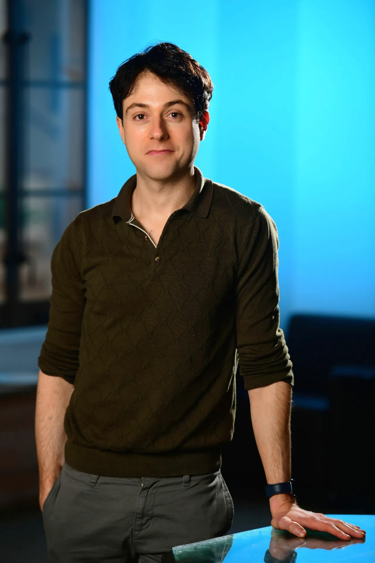 A person with short dark hair wearing a dark green sweater and gray pants stands indoors, resting one hand on a glass surface&mdash;perhaps contemplating ways to cure coma. The background is softly lit with blue tones.