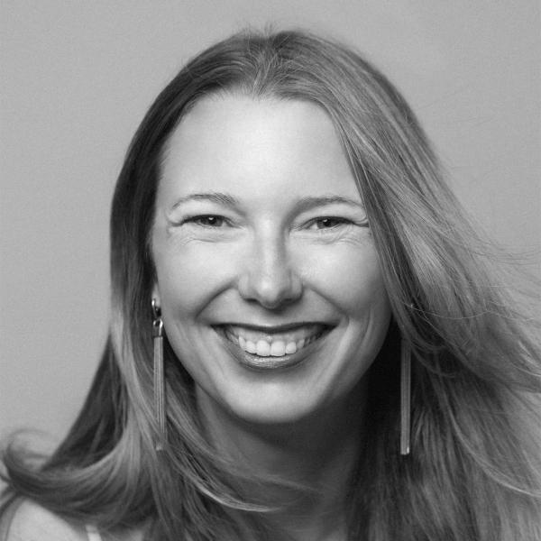 A woman with long hair smiles at the camera. She is wearing long earrings and a light-colored top. The photo is in black and white.