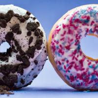 Two donuts with white icing on a blue background. The donut on the left is topped with cookie crumbles, and the one on the right is decorated with pink and blue sprinkles.