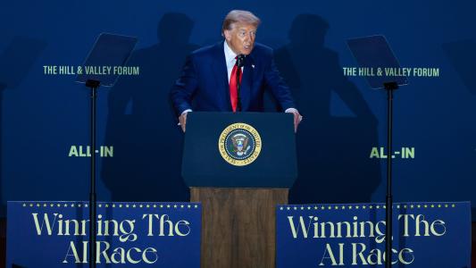 A man in a suit stands at a podium with the presidential seal, speaking at an event with signs that read "Winning The AI Race" and "ALL-IN.