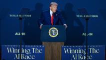 A man in a suit stands at a podium with the presidential seal, speaking at an event with signs that read "Winning The AI Race" and "ALL-IN.