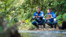 Two people in blue shirts and safety glasses analyze data with electronic devices near a stream in a forested area, exemplifying the growing field of green jobs focused on environmental conservation.