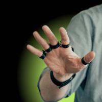 A person wearing a gray shirt is extending their hand with rings on each finger against a dark background with a green glow.