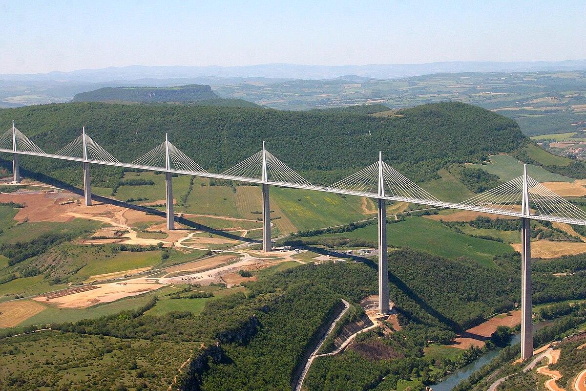 A long cable-stayed bridge with multiple pylons spans a wide valley, surrounded by green hills and farmland under a clear sky.