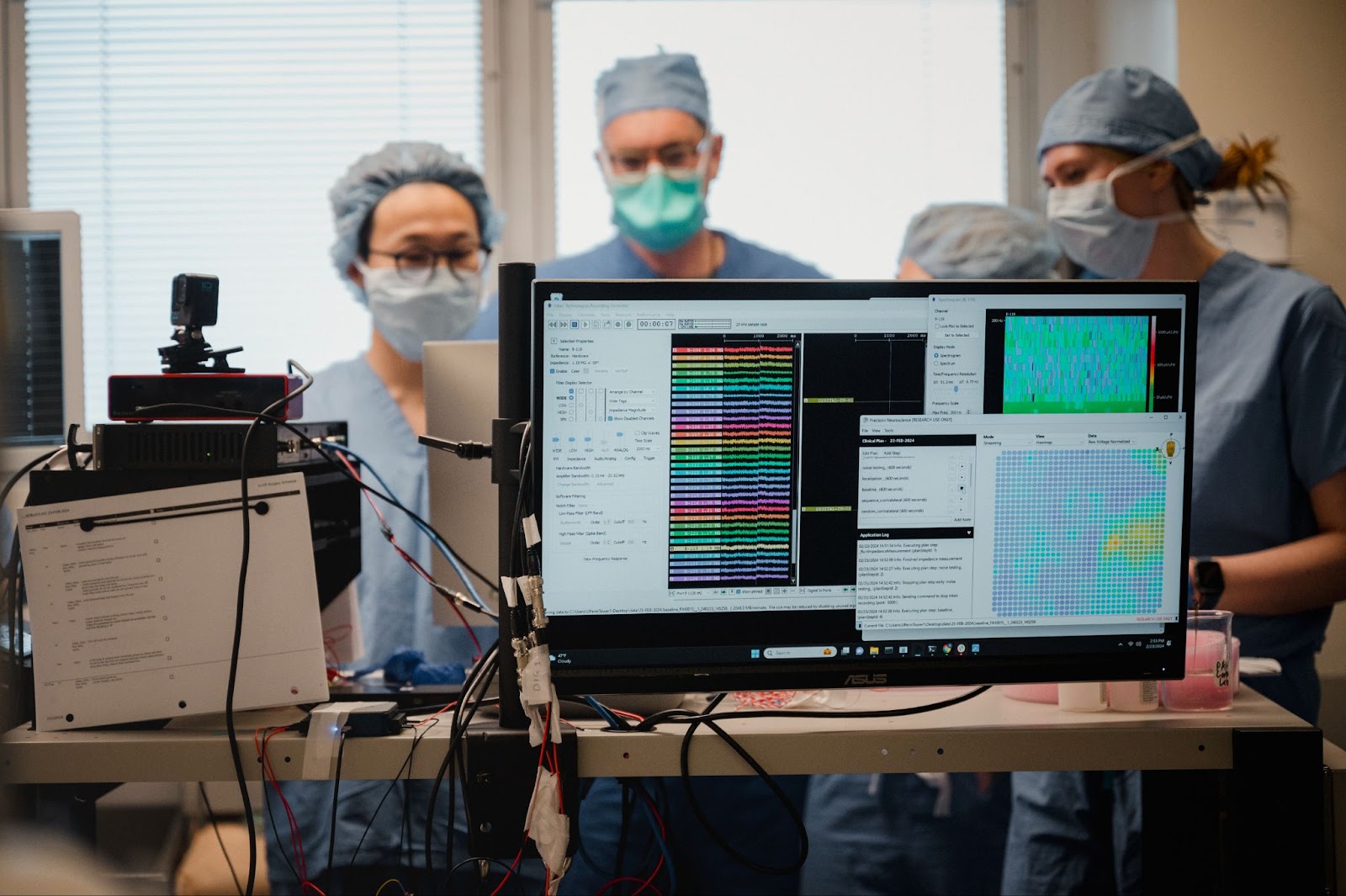 Several medical professionals in surgical scrubs and masks stand behind a monitor displaying colorful brainwave data and analysis graphs in a clinical setting.