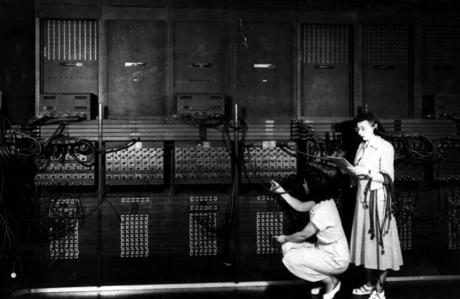 Two women operate the ENIAC computer, connecting cables and checking notes in front of a large panel filled with switches, wires, and machinery.