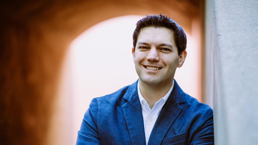 Man in a blue jacket and white shirt smiling with arms crossed, leaning against a wall, with an arched, blurred background.