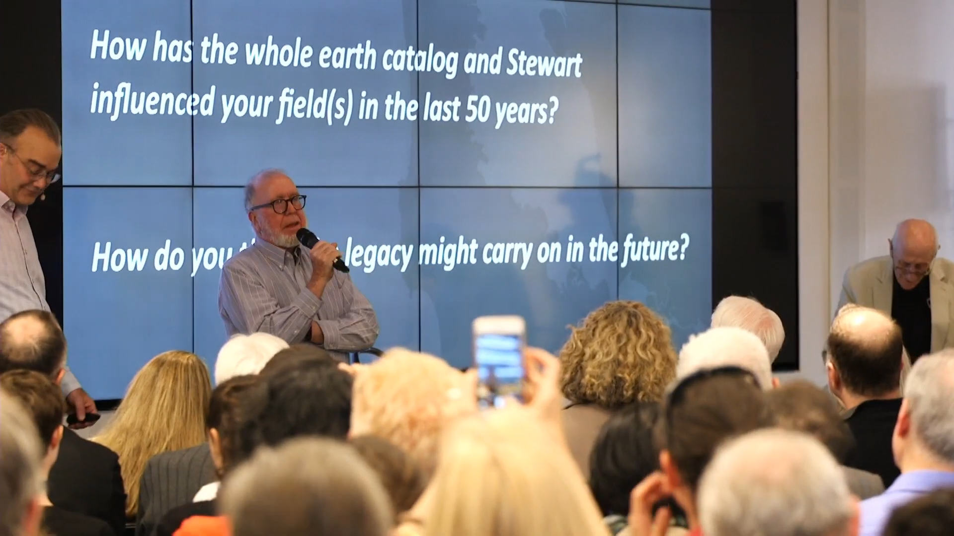 A man speaks into a microphone in front of a large audience. A screen displays questions about the impact of the Whole Earth Catalog. People hold up phones to capture the moment.
