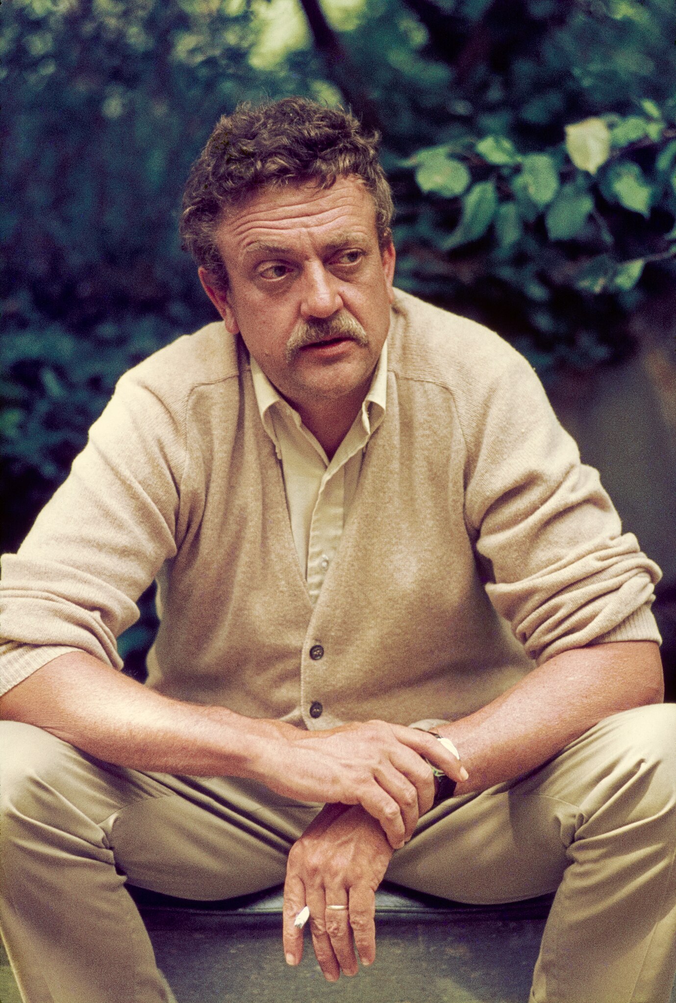 A middle-aged man with curly hair and a mustache sits outdoors, wearing a beige cardigan and holding a cigarette, with greenery in the background.