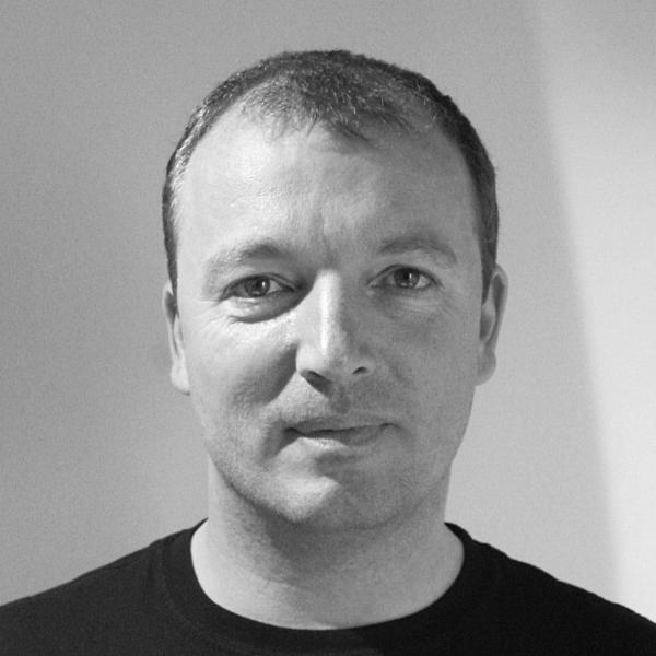 A man with short hair and a neutral expression is facing the camera against a plain, light-colored background.