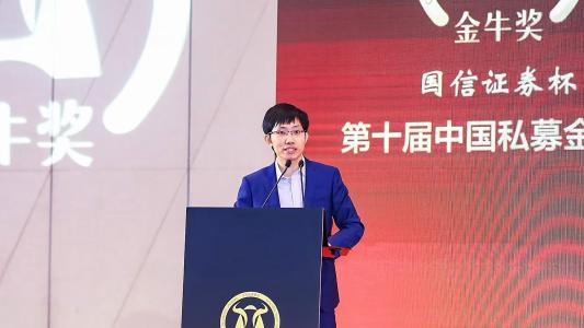 A person in a blue suit speaks at a podium during an event, with a large display screen in the background featuring Chinese text.
