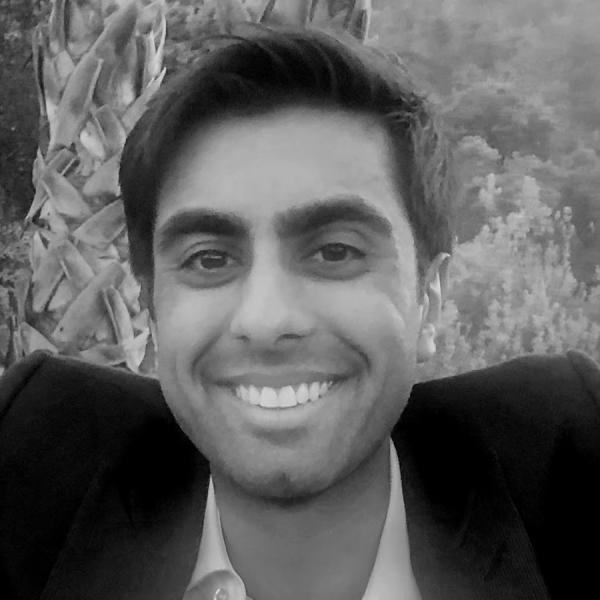 A man in a suit smiles at the camera with trees and a large plant in the background. The image is in black and white.