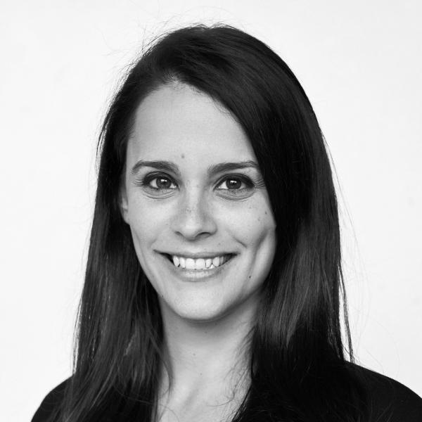 Woman with long dark hair smiling, wearing a dark top, facing the camera against a plain light background.