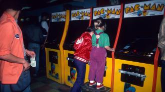 Two children play Pac-Man arcade machines in a dimly lit arcade. One child stands on a stool, while an adult nearby holds cash.