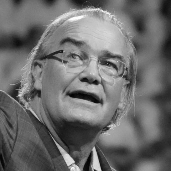 Person with glasses speaking, wearing a suit jacket, captured in black and white.