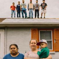 Two women and a young boy stand in front of a house while five individuals stand on the roof in the background.