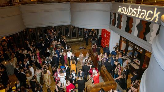 A crowd of people in formal attire socialize at an indoor event with a large "Substack" sign illuminated on the wall above.