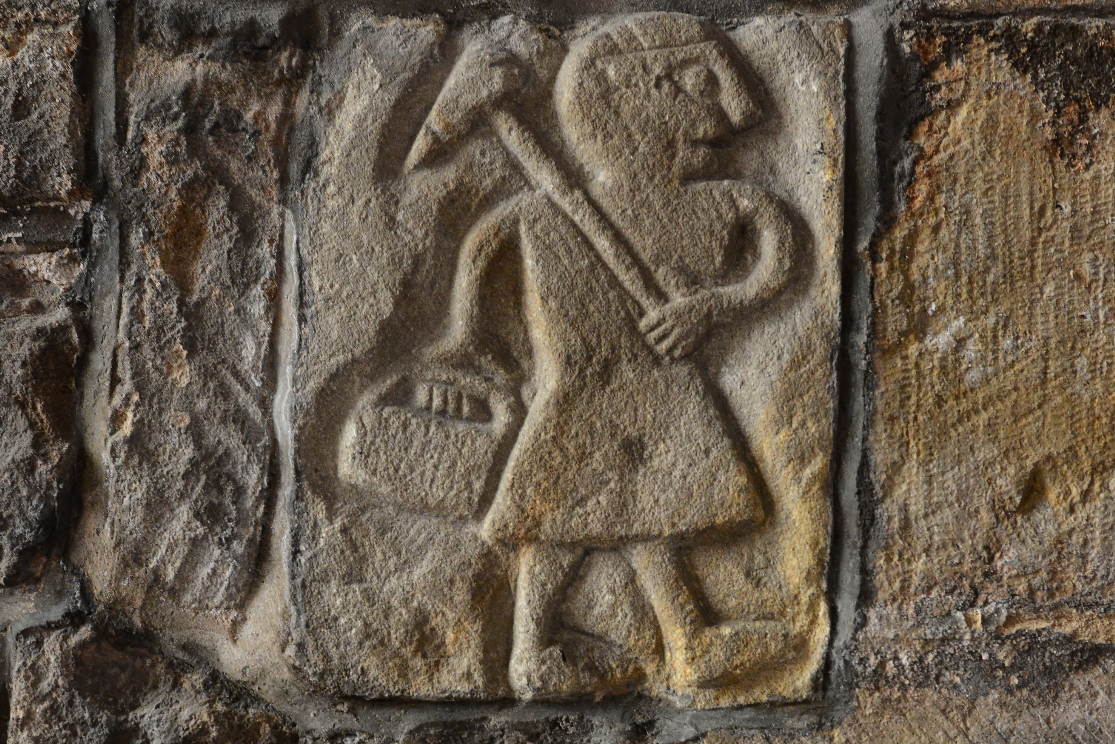 Stone carving of a person in a tunic carrying a tool over their shoulder and holding a basket, set into a rough stone wall.