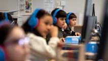 Four children wearing blue headphones use desktop computers in a classroom, focusing on their screens.