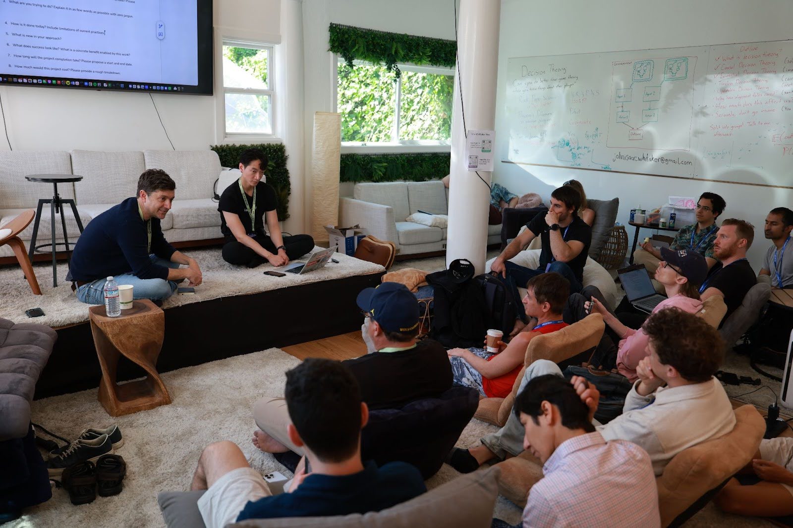 A group of people sits in a casual, modern room, listening to two presenters seated at the front near a screen and a whiteboard filled with notes.
