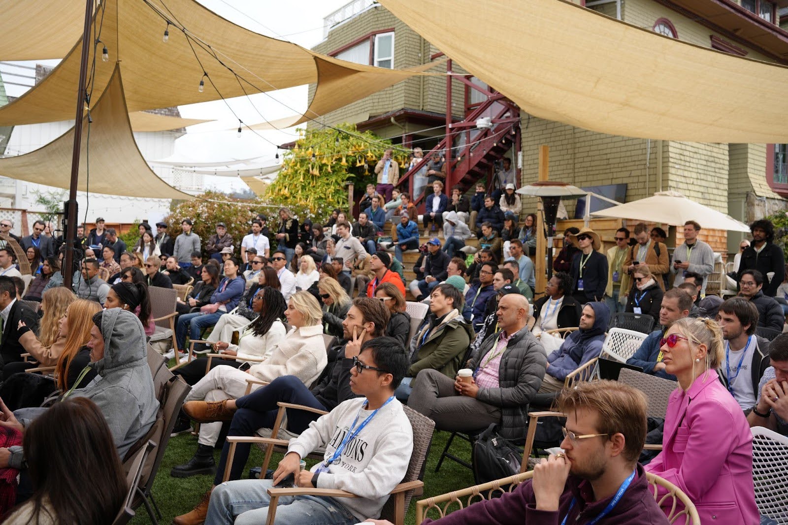 A large group of people sits outdoors under beige fabric canopies, attentively watching an event or presentation in a garden area with a house in the background.