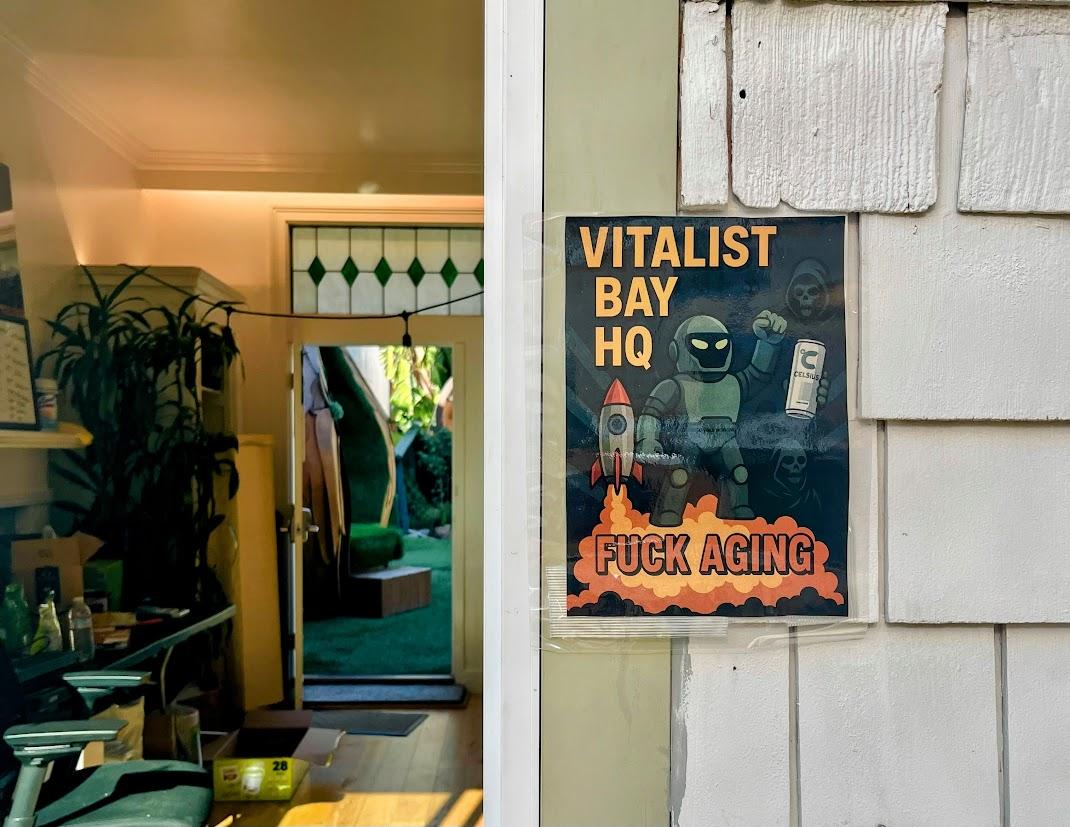 Poster on a wall reads &ldquo;Vitalist Bay HQ Fuck Aging&rdquo; with cartoon figures, a rocket, and lab items; an open door reveals a room with plants and green decor inside.