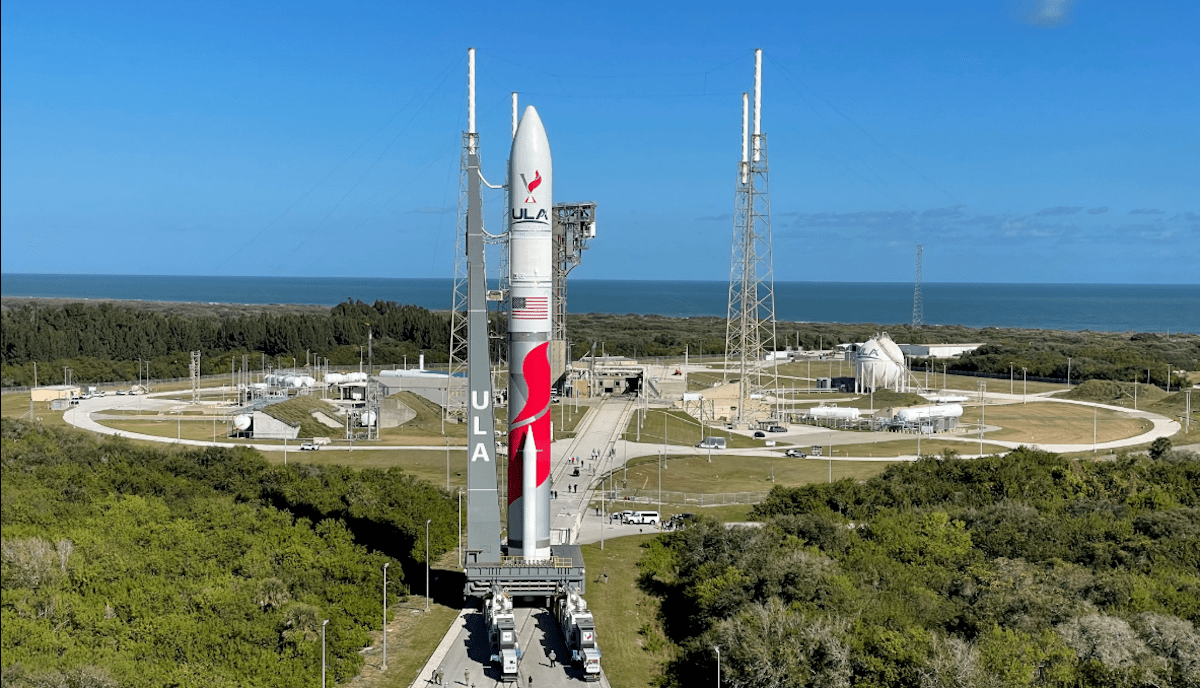 A large rocket is positioned vertically on a launch pad, surrounded by green foliage and set against a clear blue sky. Structures and equipment are visible in the background.