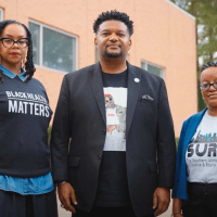 Three people stand outside a brick building, facing forward. The woman on the left wears a "Black Health Matters" shirt, the man in the center wears a blazer, and the woman on the right wears glasses and a blue blazer.