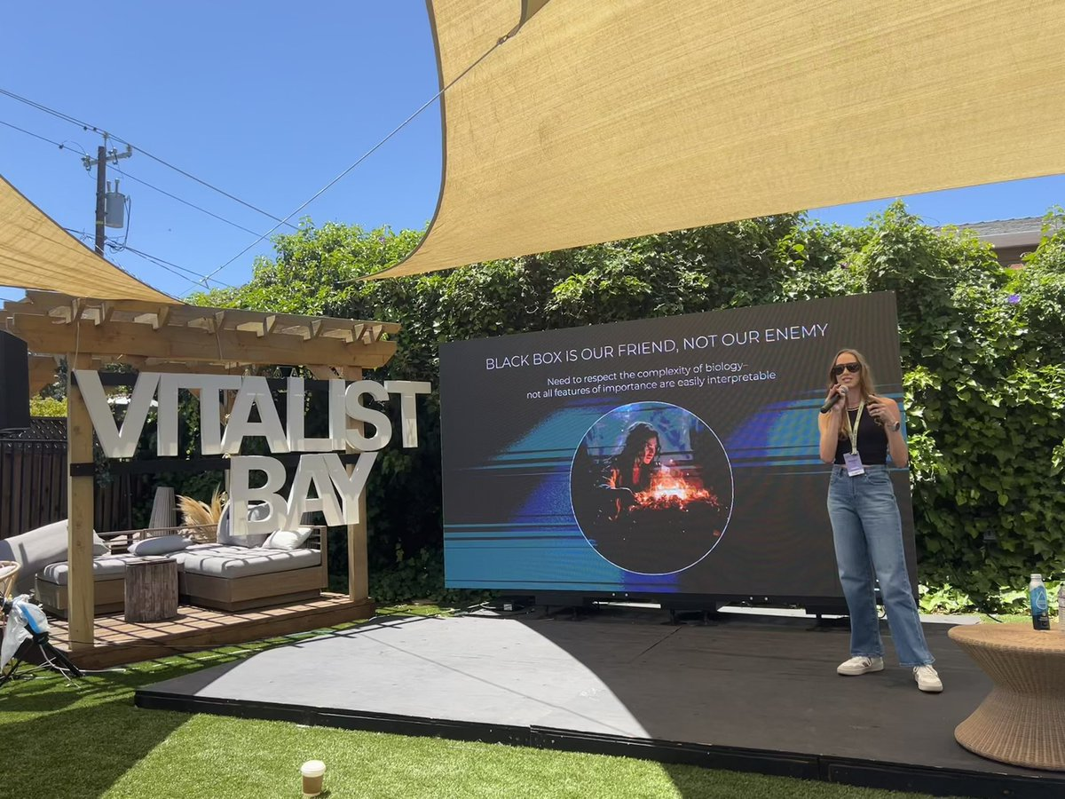 A woman speaks on an outdoor stage with a screen displaying a presentation titled "BLACK BOX IS OUR FRIEND, NOT OUR ENEMY" at an event with "VITALIST BAY" sign in the background.